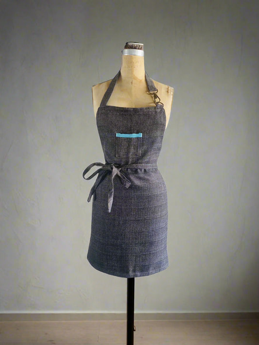 A apron with a solid side and aqua colored rim detail, featuring a double-sided, round edges design with a front pocket on the solid side, a solid fabric apron strap that ties around the waist, a snap hook detail on one side of the apron strap.