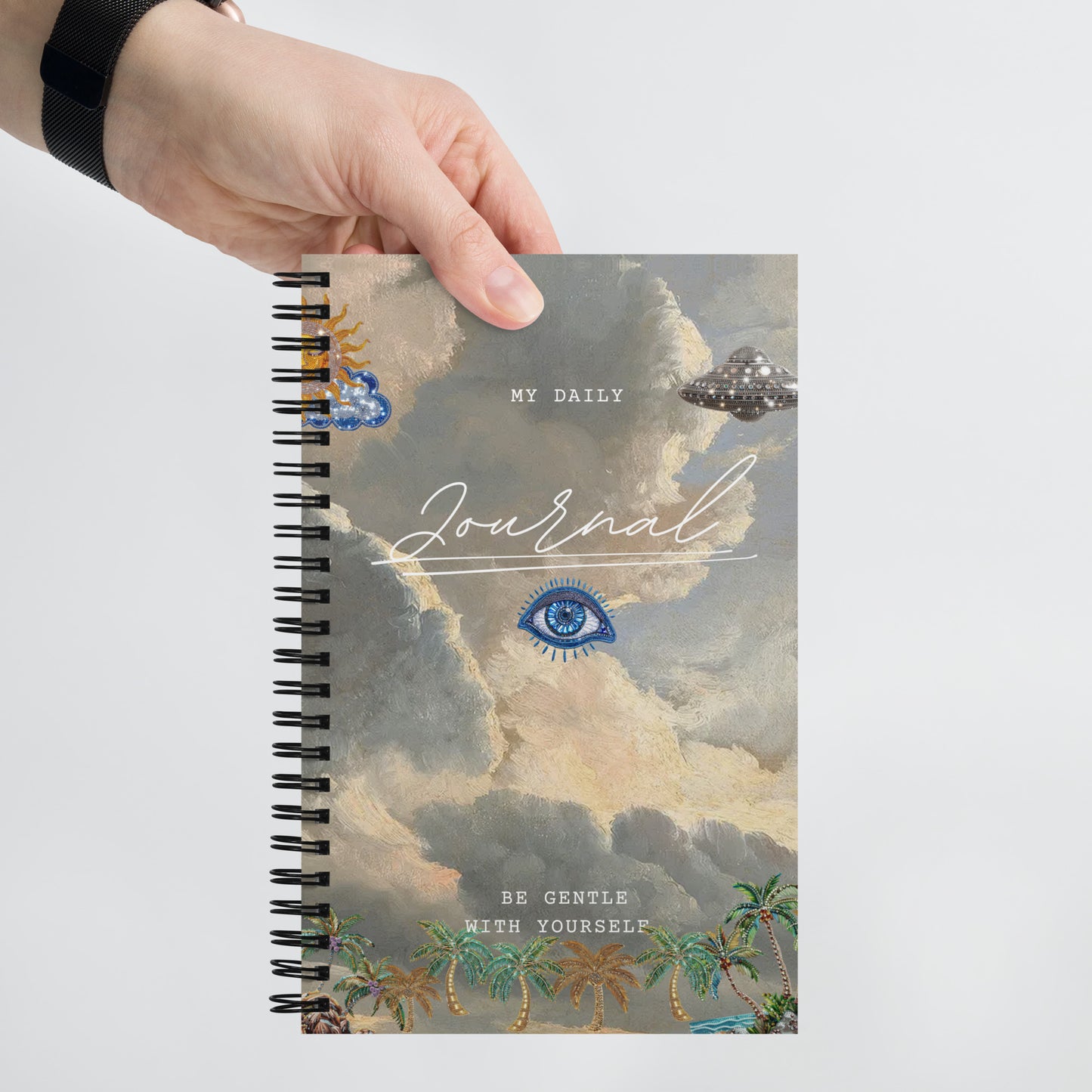 My Daily Journal spiral notebook held in hands, showcasing detail of front art cover that includes eye design, clouds, gold accents, and palm trees, with 'Be Gentle With Yourself' text