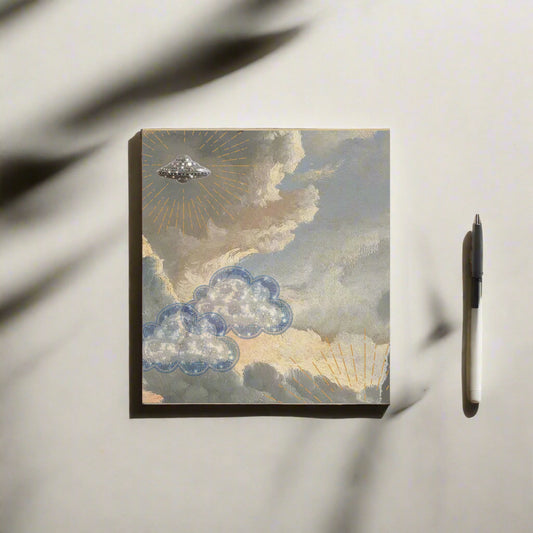 Cloud notepad with sequined blue clouds and UFO on dreamy sky background laying on top of table with a pen ready to start jotting down ideas