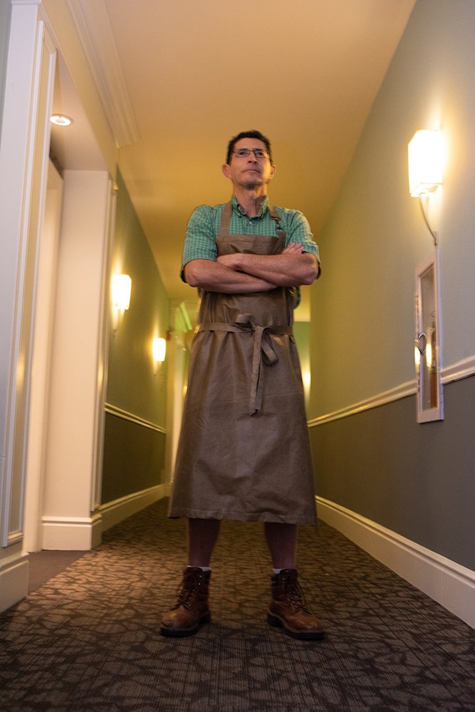 Chef in sage green shirt and charcoal apron standing in modern hallway